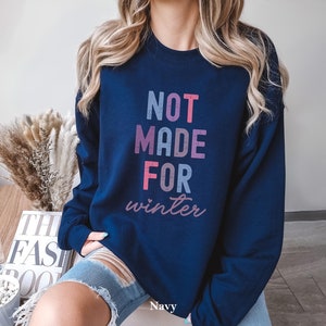 May include: A navy blue sweatshirt with the text "NOT MADE FOR winter" in pink, blue, and purple lettering. The sweatshirt is being worn by a person with long blonde hair.