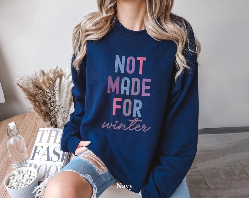 May include: A navy blue sweatshirt with the text "NOT MADE FOR winter" in pink, blue, and purple lettering. The sweatshirt is being worn by a person with long blonde hair.