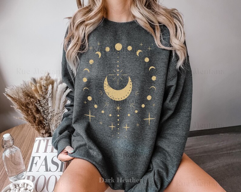 以下が含まれることがあります： ダークヘザーグレーのトレーナーに、金の月と星のグラフィックがプリントされています。グラフィックは、星が内側に描かれた三日月と、その周りを囲む小さな月と星の円を描いています。画像の下部には「ダークヘザー」というテキストが表示されています。