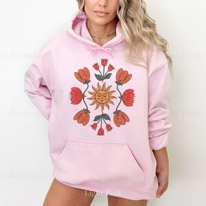 May include: A light pink hoodie with a graphic print of a sun with a face and flowers in shades of orange, red, and green.