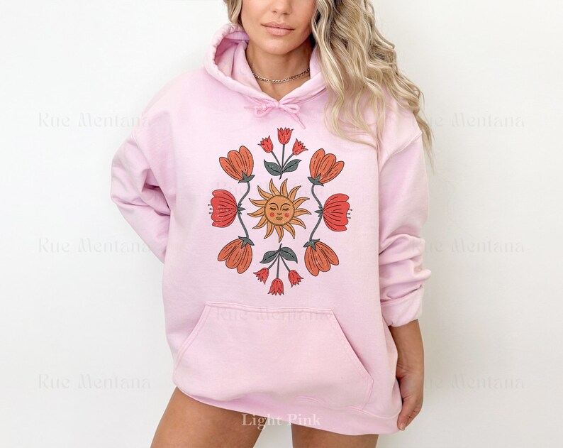 May include: A light pink hoodie with a graphic print of a sun with a face and flowers in shades of orange, red, and green.