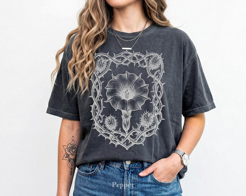 Puede incluir: Camiseta gris oscuro con un dise&ntilde;o floral blanco detallado dentro de un marco espinoso. El dise&ntilde;o incluye una flor grande y elementos similares a cardos. La camiseta es de corte holgado, con un lavado vintage.