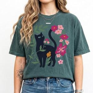 Pu&ograve; includere: Una t-shirt a maniche corte color verde acqua scuro con una stampa grafica di un gatto nero circondato da fogliame verde e vari fiori rosa e magenta. Il gatto &egrave; posizionato centralmente con la coda rivolta verso l'alto e un piccolo baccello arancione &egrave; visibile.