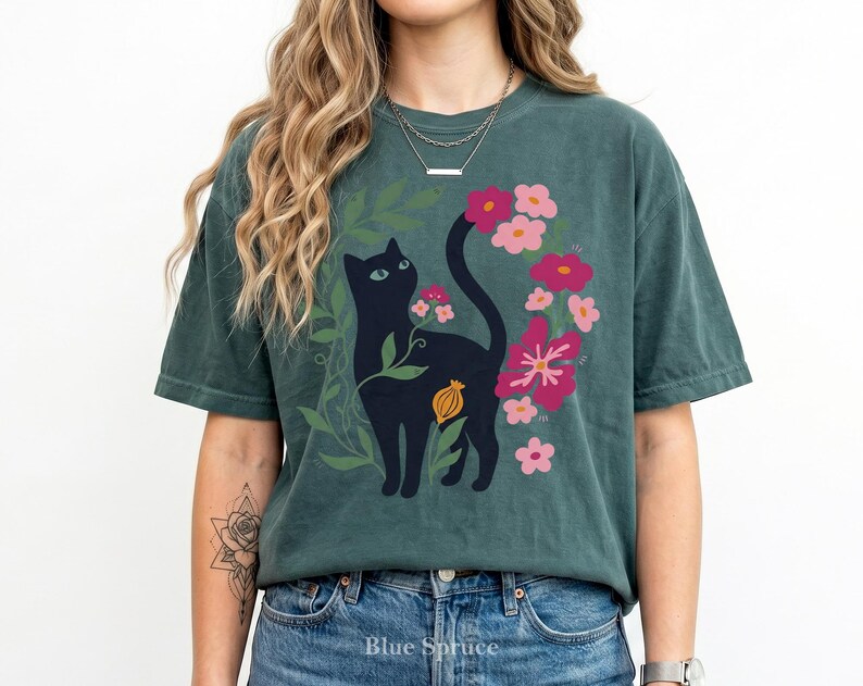 Pu&ograve; includere: Una t-shirt a maniche corte color verde acqua scuro con una stampa grafica di un gatto nero circondato da fogliame verde e vari fiori rosa e magenta. Il gatto &egrave; posizionato centralmente con la coda rivolta verso l'alto e un piccolo baccello arancione &egrave; visibile.