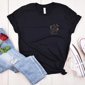 K&ouml;nnte beinhalten: Schwarzes T-Shirt mit einer goldenen Illustration einer Frau, eines Mondes und Bl&auml;ttern. Das Shirt wird mit hellblauen Jeans, einer roten Rose und wei&szlig;en Turnschuhen mit rot-blauen Streifen kombiniert.