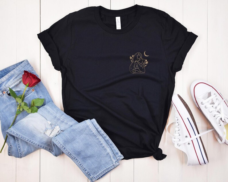 K&ouml;nnte beinhalten: Schwarzes T-Shirt mit einer goldenen Illustration einer Frau, eines Mondes und Bl&auml;ttern. Das Shirt wird mit hellblauen Jeans, einer roten Rose und wei&szlig;en Turnschuhen mit rot-blauen Streifen kombiniert.