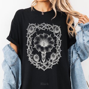 Puede incluir: Camiseta negra con un dise&ntilde;o floral blanco detallado dentro de un marco espinoso. El dise&ntilde;o incluye una flor grande y capullos m&aacute;s peque&ntilde;os. La palabra "Black" est&aacute; impresa en la parte inferior de la camiseta. La camiseta tiene mangas cortas.