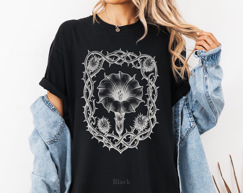Puede incluir: Camiseta negra con un dise&ntilde;o floral blanco detallado dentro de un marco espinoso. El dise&ntilde;o incluye una flor grande y capullos m&aacute;s peque&ntilde;os. La palabra "Black" est&aacute; impresa en la parte inferior de la camiseta. La camiseta tiene mangas cortas.