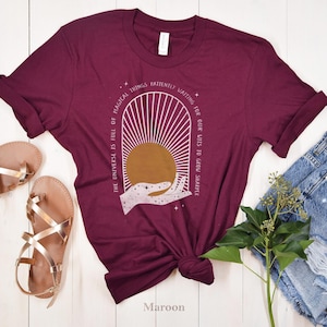 May include: Maroon t-shirt with a white graphic design of a hand holding a sun with the text "The universe is full of magical things patiently waiting for our wits to grow sharper".
