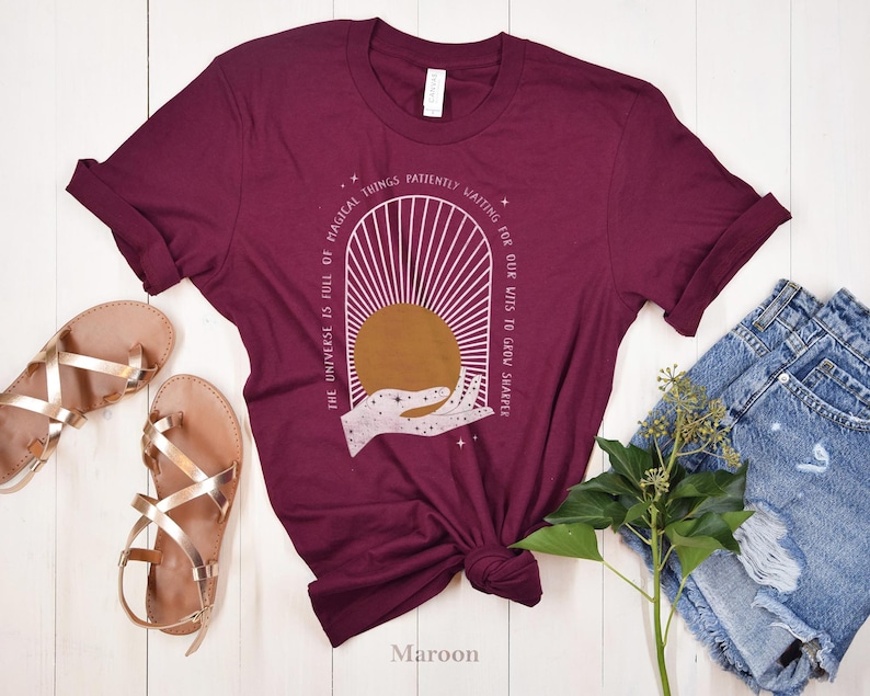 May include: Maroon t-shirt with a white graphic design of a hand holding a sun with the text "The universe is full of magical things patiently waiting for our wits to grow sharper".