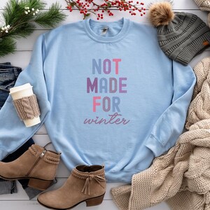 May include: A light blue sweatshirt with the text "NOT MADE FOR winter" in pink, purple, and blue lettering. The sweatshirt is laid out on a white wooden surface with a pair of brown ankle boots, a pair of blue jeans, a knitted scarf, and a gray beanie with a brown pom-pom.