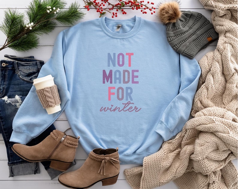 May include: A light blue sweatshirt with the text "NOT MADE FOR winter" in pink, purple, and blue lettering. The sweatshirt is laid out on a white wooden surface with a pair of brown ankle boots, a pair of blue jeans, a knitted scarf, and a gray beanie with a brown pom-pom.