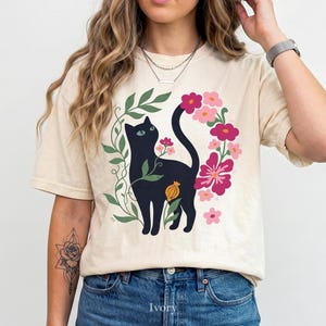 Pu&ograve; includere: Una t-shirt a maniche corte color avorio con una stampa grafica di un gatto nero circondato da viti verdi e vari fiori rosa e magenta. Il gatto &egrave; posizionato centralmente, con la coda curva verso l'alto tra gli elementi floreali.
