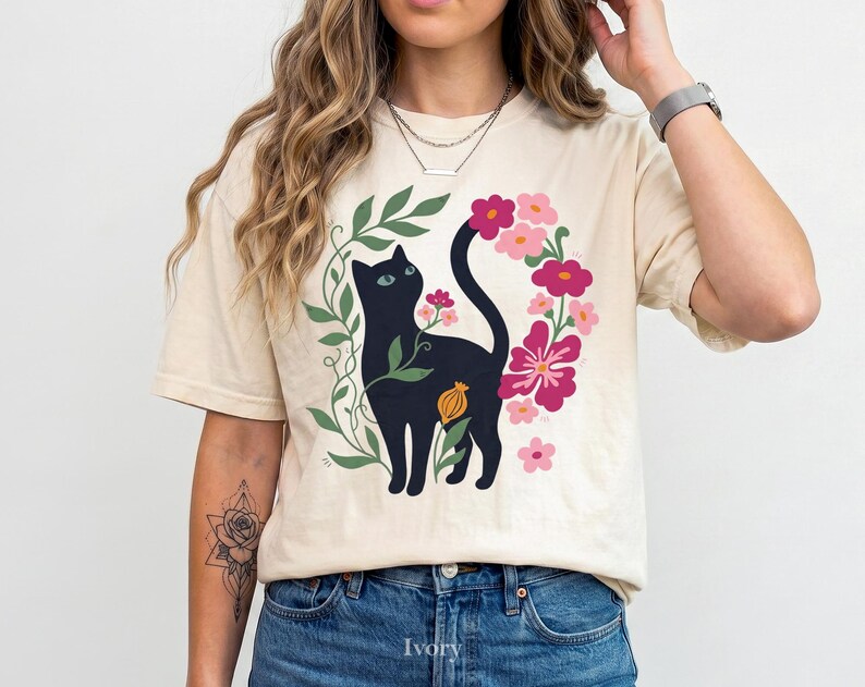 Pu&ograve; includere: Una t-shirt a maniche corte color avorio con una stampa grafica di un gatto nero circondato da viti verdi e vari fiori rosa e magenta. Il gatto &egrave; posizionato centralmente, con la coda curva verso l'alto tra gli elementi floreali.