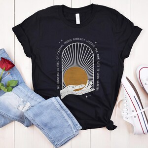 May include: Black t-shirt with a white graphic design of a hand reaching towards a sun with rays. The text "The universe is full of magical things patiently waiting for our wits to grow sharper" is printed on the shirt.