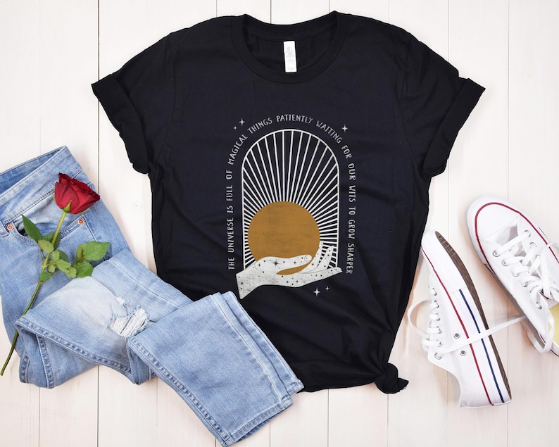 May include: Black t-shirt with a white graphic design of a hand reaching towards a sun with rays. The text "The universe is full of magical things patiently waiting for our wits to grow sharper" is printed on the shirt.