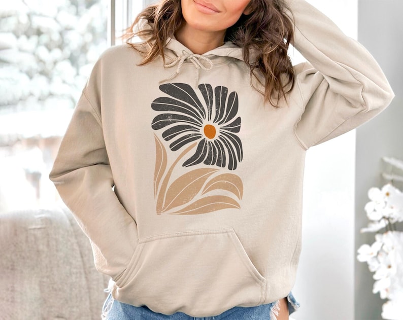 K&ouml;nnte beinhalten: Ein beiger Hoodie mit einem schwarzen und braunen Blumendesign. Die Blume hat eine gelbe Mitte.