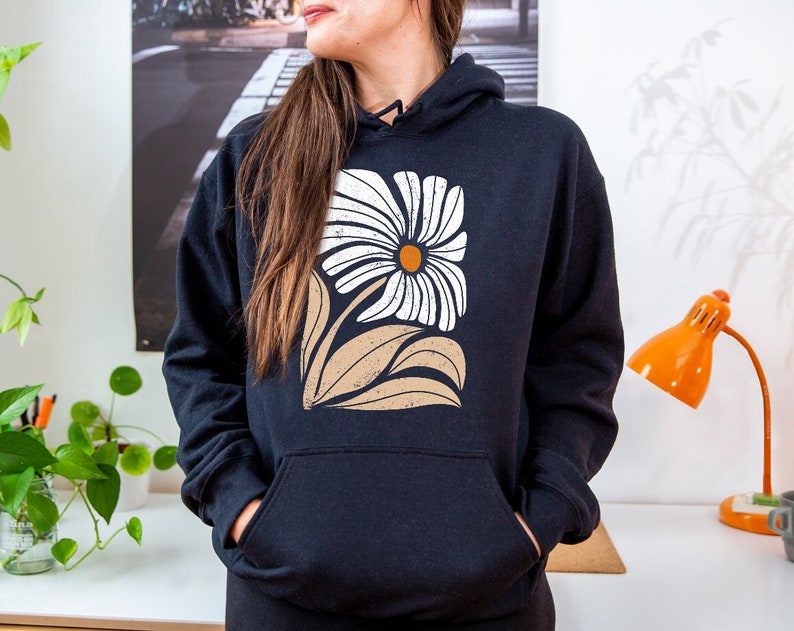 K&ouml;nnte beinhalten: Ein schwarzer Hoodie mit einem wei&szlig;en G&auml;nsebl&uuml;mchen-Druck. Die Blume hat eine braune Mitte und ist von braunen Bl&auml;ttern umgeben.