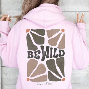 Op de afbeelding: Licht roze hoodie met een bruin en beige abstract ontwerp en de tekst "BE WILD" op de rug.