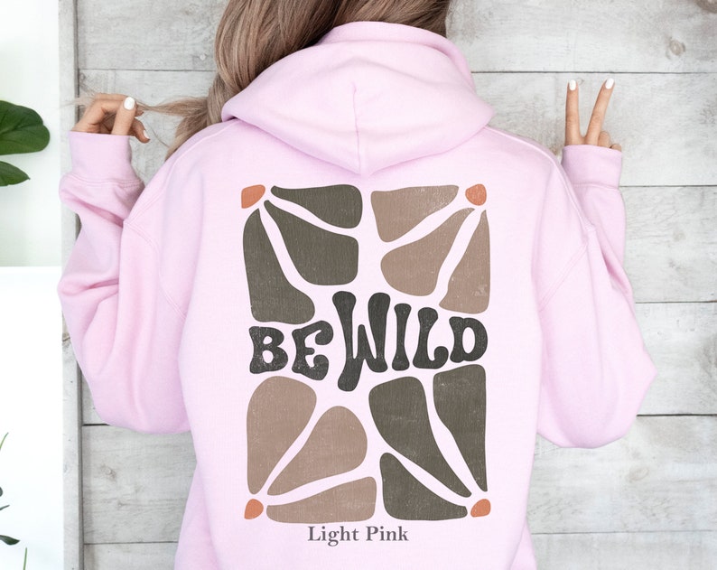 Op de afbeelding: Licht roze hoodie met een bruin en beige abstract ontwerp en de tekst "BE WILD" op de rug.