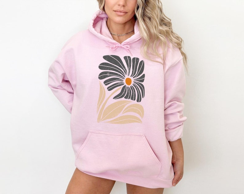 K&ouml;nnte beinhalten: Ein rosa Hoodie mit einem schwarzen und beigefarbenen Blumendesign. Die Blume hat eine gro&szlig;e, dunkle Mitte mit einem kleinen orangefarbenen Kreis in der Mitte.