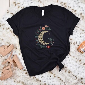 Może przedstawiać: Czarny t-shirt z motywem p&oacute;łksiężyca otoczonym złotymi, brzoskwiniowymi i czerwonymi kwiatami i liśćmi. Na zdjęciu znajdują się r&oacute;wnież sandały na platformie i butelka wina musującego.