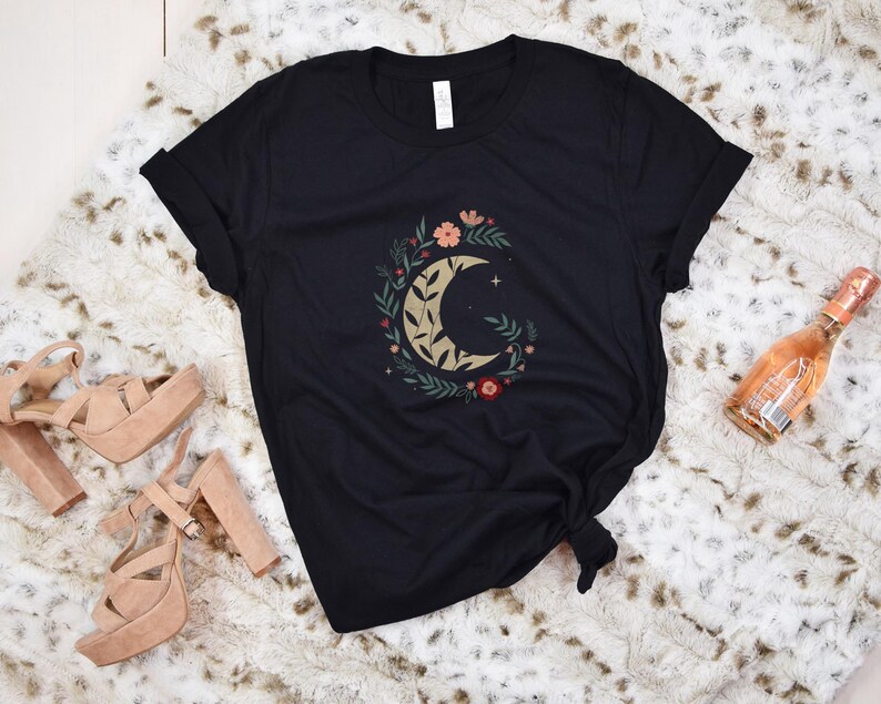 Może przedstawiać: Czarny t-shirt z motywem p&oacute;łksiężyca otoczonym złotymi, brzoskwiniowymi i czerwonymi kwiatami i liśćmi. Na zdjęciu znajdują się r&oacute;wnież sandały na platformie i butelka wina musującego.