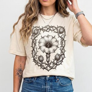 Puede incluir: Camiseta color crema con una ilustraci&oacute;n floral en blanco y negro. El dise&ntilde;o presenta una gran flor enmarcada por espinas y c&aacute;psulas de semillas puntiagudas. La camiseta est&aacute; hecha de un material suave y tiene un corte holgado.
