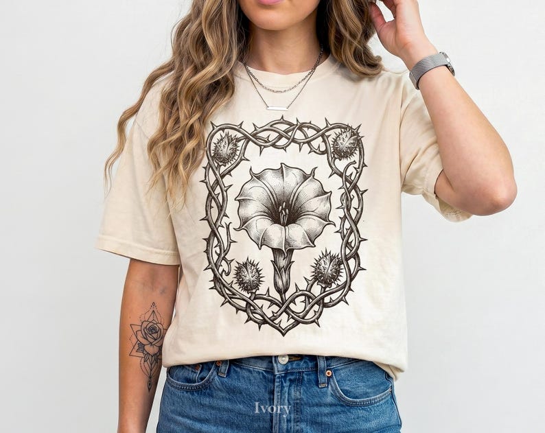 Puede incluir: Camiseta color crema con una ilustraci&oacute;n floral en blanco y negro. El dise&ntilde;o presenta una gran flor enmarcada por espinas y c&aacute;psulas de semillas puntiagudas. La camiseta est&aacute; hecha de un material suave y tiene un corte holgado.