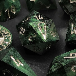 Könnte beinhalten: Eine Sammlung grüner, polyedrischer Würfel mit silbernen Zahlen und Markierungen. Die Würfel haben verschiedene geometrische Formen. Ein kreisförmiges Token mit Wolfsmotiv ist ebenfalls vorhanden.