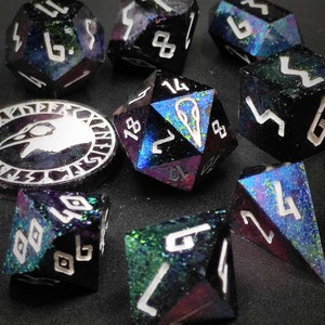 Puede incluir: Un conjunto de dados de diez caras con pintura negra y verde iridiscente y números blancos. Los dados están sobre una superficie negra con una moneda de plata que presenta un diseño de cuervo.