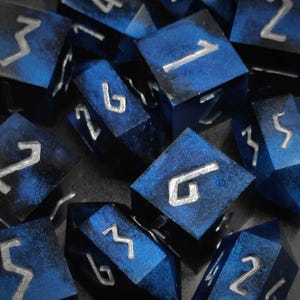 Puede incluir: Un conjunto de dados azules y plateados con un patrón de galaxia. Los dados están numerados del 1 al 6.