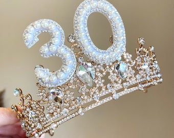 Può includere: Corona dorata con strass trasparenti e bianchi, con un numero "30" incrostato di perle bianche in cima.