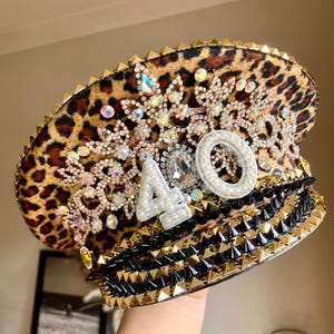 May include: A leopard print captain's hat with gold and black spikes, rhinestones, and the number 40. The hat is heavily embellished with sparkling jewels and pearls. The brim is lined with gold triangles.