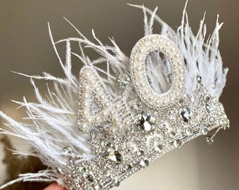 May include: A silver-toned crown with the number "40" embellished with pearls and crystals. White feathers accent the crown's design, adding a touch of elegance. The crown is likely for a special occasion.