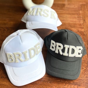 Może przedstawiać: Trzy białe i czarne siatkowe czapki trucker z perłowymi ozdobami. Czapki mają napis "MRS", "BRIDE" i "BRIDE".