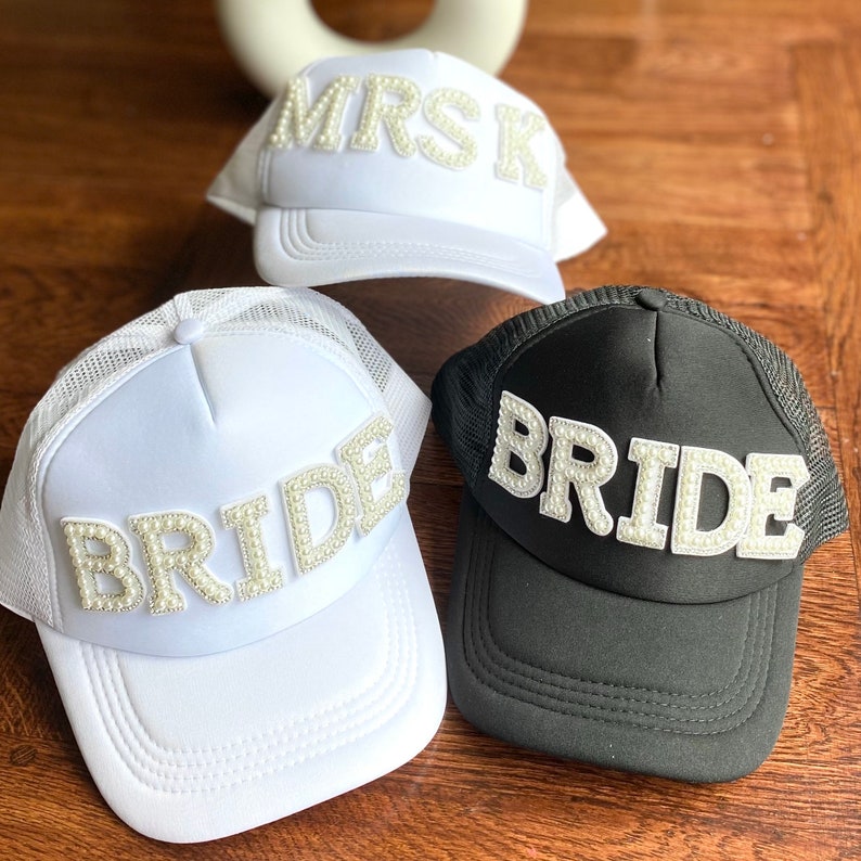 Może przedstawiać: Trzy białe i czarne siatkowe czapki trucker z perłowymi ozdobami. Czapki mają napis "MRS", "BRIDE" i "BRIDE".