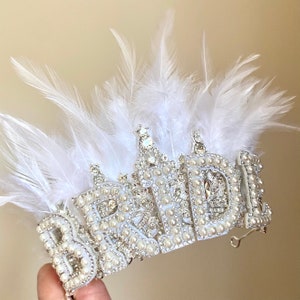 May include: White feather bridal crown with the word "BRIDE" spelled out in silver with pearl accents.
