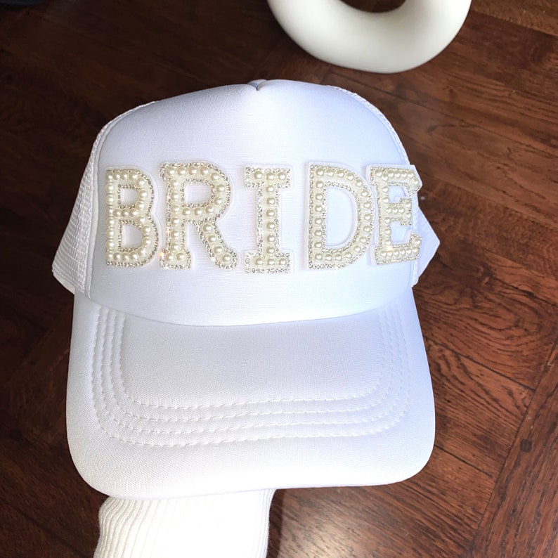 Może przedstawiać: Biała siatkowa czapka trucker z białym, ozdobionym perłami napisem "BRIDE".