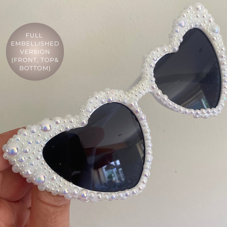 May include: White heart-shaped sunglasses with black lenses, fully embellished with iridescent pearls.