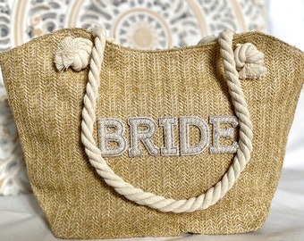 May include: A beige straw tote bag with white rope handles and the word "BRIDE" in white letters with pearl accents.