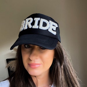 Może przedstawiać: Czarna siatkowa czapka trucker z białym, ozdobionym perłami napisem "BRIDE". Czapka ma zakrzywiony daszek i regulowany pasek.