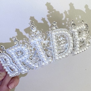 May include: A white bridal crown with the word "BRIDE" spelled out in silver letters and decorated with white pearls.