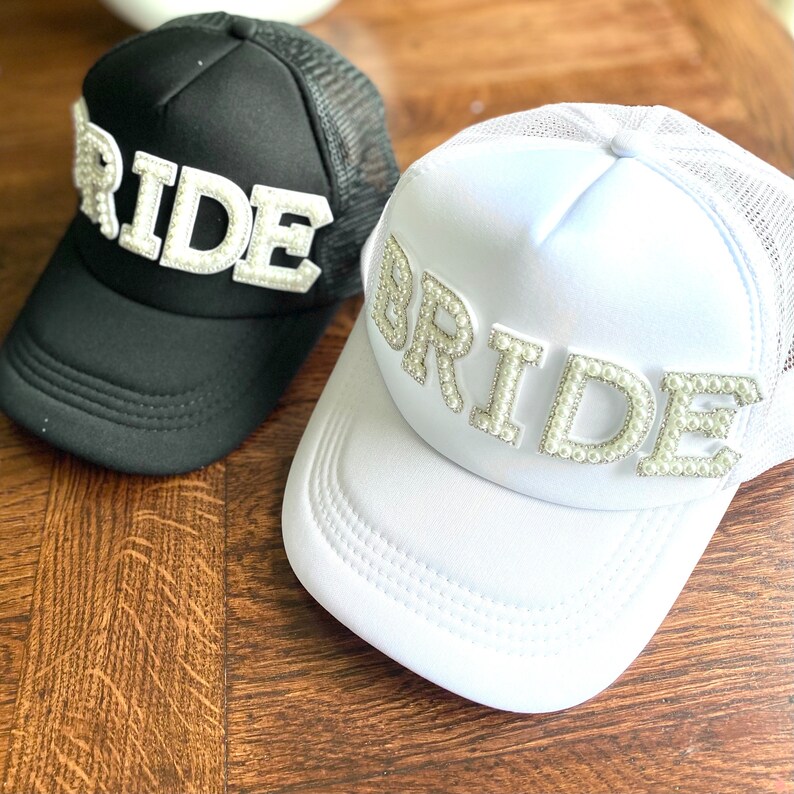 Może przedstawiać: Dwie czapki trucker, jedna czarna, a druga biała, z napisem "BRIDE" napisanym białymi perłami. Czarna czapka ma napis "RIDE" białymi literami.