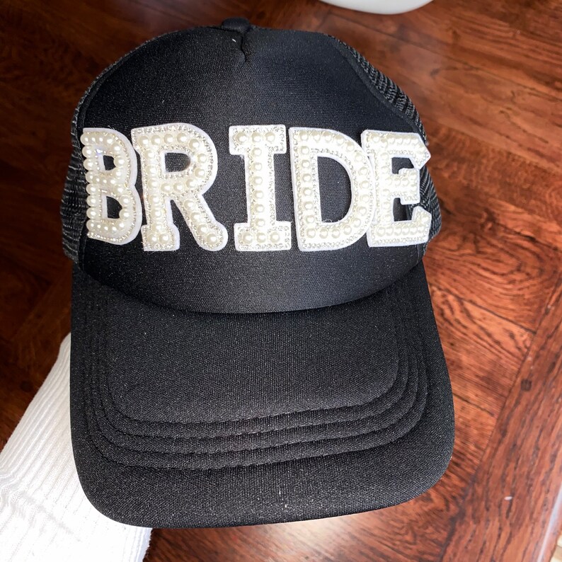 Może przedstawiać: Czarna czapka trucker z białym napisem "BRIDE" ozdobionym koralikami imitującymi perły.
