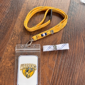 Peut inclure: Un cordon jaune avec des points noirs et un fermoir argenté, un porte-badge en plastique transparent avec le logo "Warriors", et un morceau de lacet blanc avec le chiffre "9" en bleu. Les articles sont sur une surface en bois.