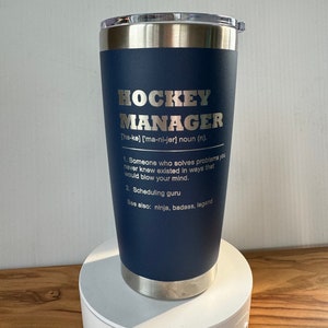 May include: A blue stainless steel tumbler with a silver lid. The tumbler has a white inscription that reads "HOCKEY MANAGER ['ha-ke] ['ma-ni-jer] noun (n). 1. Someone who solves problems you never knew existed in ways that would blow your mind. 2. Scheduling guru See also: ninja, badass, legend"