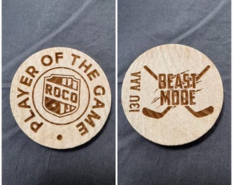 May include: Two circular light-colored wooden coasters. The left coaster features a shield graphic with the text ROCO and the words PLAYER OF THE GAME. The right coaster displays two crossed hockey sticks with the text 13U AAA BEAST MODE.