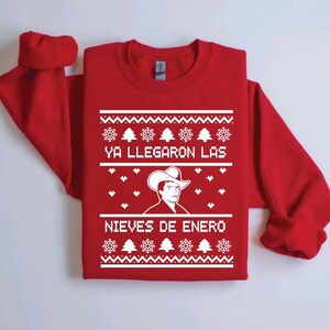 Peut inclure: Pull de Noël rouge avec un motif de flocons de neige et de sapins blancs. Le pull présente une image blanche d'un homme portant un chapeau de cowboy et le texte "Ya llegaron las nieves de enero".