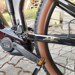 Puede incluir: Bicicleta de monta&ntilde;a el&eacute;ctrica negra con neum&aacute;tico marr&oacute;n, motor Bosch negro y plateado y pedal negro con reflector amarillo. La bicicleta tiene un cuadro negro con una inscripci&oacute;n dorada "Limited Edition".